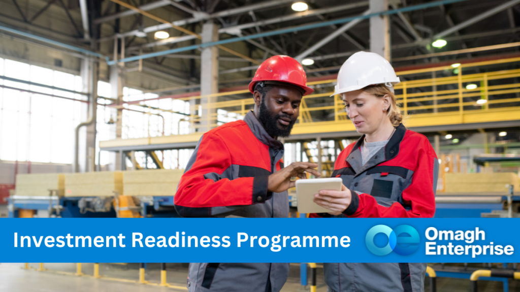 A black man, in a red body suit and red hard hat, is looking at a digital tablet with a white woman in a red boiler suit and white hard hat. They are both standing in an non-specified manufacturing factory. Blue banner along the bottom. White text "Investment Readiness Programme" Omagh Enterprise logo within the blue banner