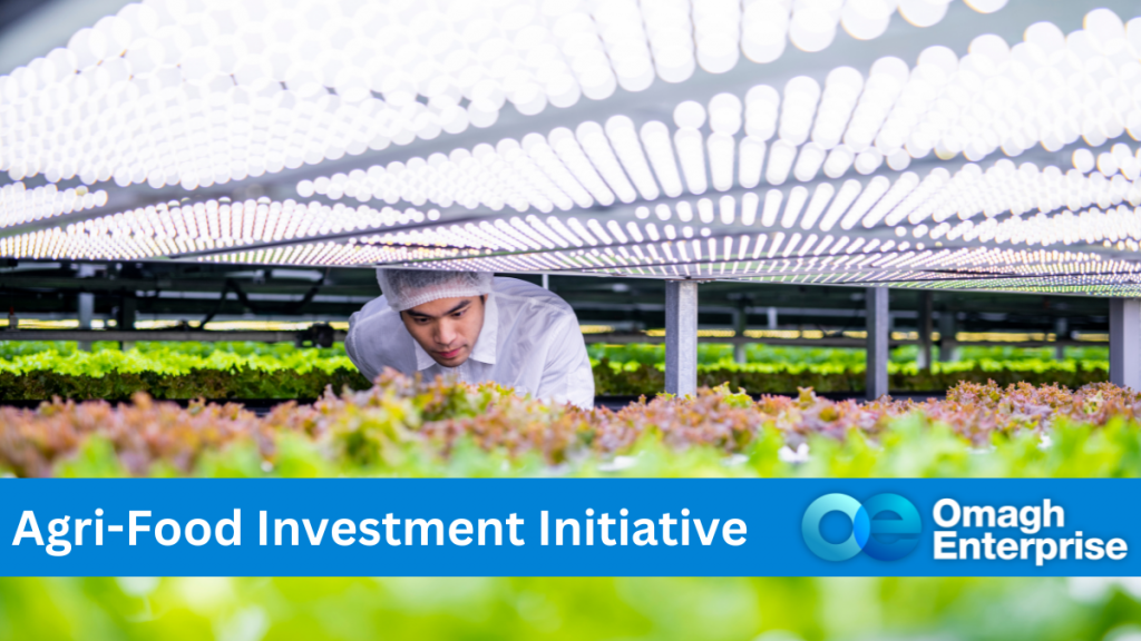 Agri-food being grown under heated lights in a factory. A man dressed in white is inspecting the food growing. Blue banner along the bottom, with white text "Agri-Food Investment Initiative" Omagh Enterprise logo within the blue banner.