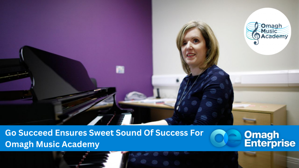 Claire Bowes is dressed in black, playing a black grand piano in her music academy. The wall behind her is purple, with vertical blinds on the window. Blue banner along the bottom, with white text. "Go Succeed Ensures Sweet Sound Of Success For Omagh Music Academy" Omagh Enterprise logo within the banner.