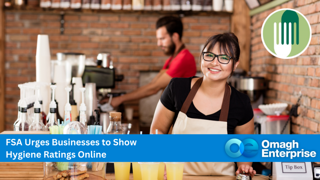 A woman behind a cafe counter smiling, wearing a black t-shirt and white apron. A man in background blurred, wearing a red t-shirt, pouring coffee from a machine. Blue banner along the bottom, with white text. "FSA Urges Businesses to Show Hygiene Ratings Online" Omagh Enterprise logo within the banner.