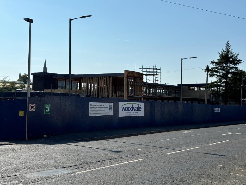 A view of the build from the Mountjoy Road. High blue fence around the site, with sign for woodvale on it. Basic brickwork shell up, with scaffolding,