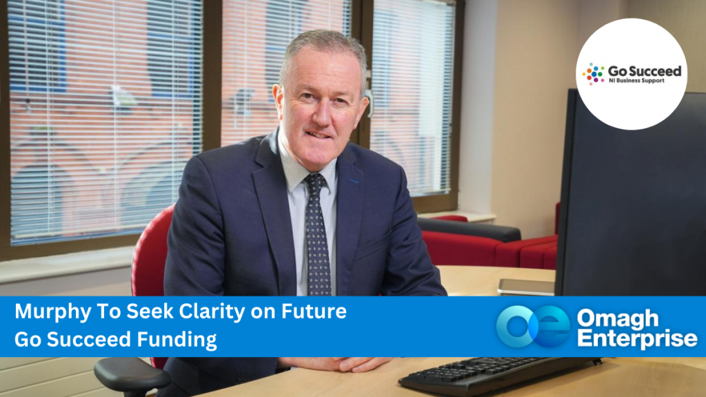 Economy Minister Conor Murphy sitting behind a desk in an office, with black computer and monitor. Caption "Murphy To Seek Clarity on Future Go Succeed Funding"