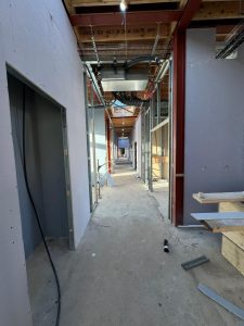 A partially constructed hallway with exposed wiring and metal framework. The walls are unfinished, and construction materials are scattered on the concrete floor. Ceiling lights illuminate the space.