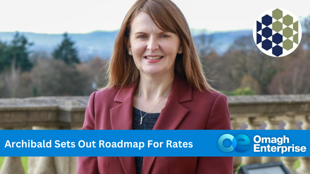 Finance Minister, Dr Caoimhe Archibald in a maroon blazer stands outside with a scenic view of trees and hills in the background from Stormont. A banner reads Archibald Sets Out Roadmap For Rates with the Omagh Enterprise logo visible.