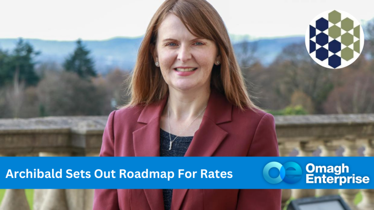 Finance Minister, Dr Caoimhe Archibald in a maroon blazer stands outside with a scenic view of trees and hills in the background from Stormont. A banner reads Archibald Sets Out Roadmap For Rates with the Omagh Enterprise logo visible.