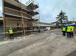 Construction site with workers in reflective vests and hard hats. A partially built structure with scaffolding is on the left. A group of people stands on the right, observing the site. Cloudy sky overhead.