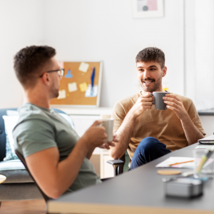 Two men sitting at a desk, holding mugs and talking in a bright office space.