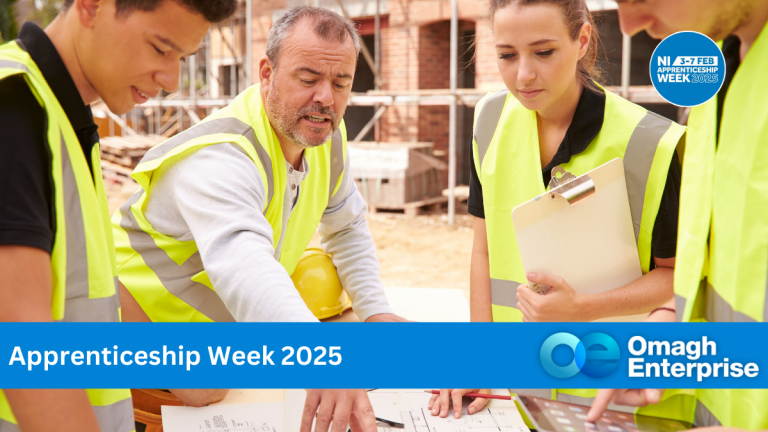 A group of four people wearing high-visibility vests gather around a construction site table reviewing plans. The text reads Apprenticeship Week 2025 and Omagh Enterprise with a logo.