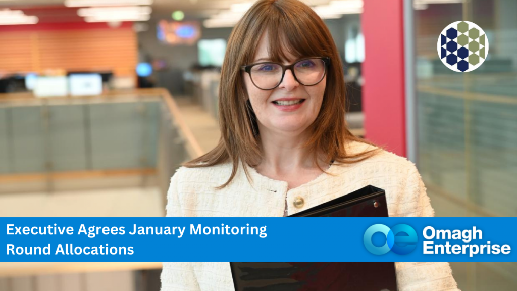 Finance Minister Dr Caoimhe Archibald with glasses smiles, holding a black folder. Text reads: "Executive Agrees January Monitoring Round Allocations - Omagh Enterprise.