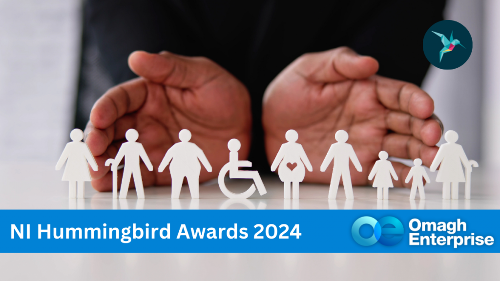 A pair of hands encloses a row of white paper cutout figures, including a person in a wheelchair, on a reflective surface. Text reads NI Hummingbird Awards 2024, with the Omagh Enterprise logo. A small bird icon is in the top right corner.