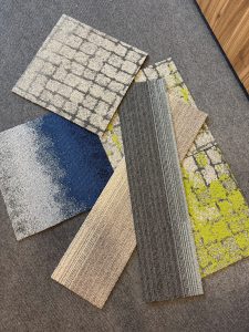 A variety of carpet samples are displayed on a gray floor. The samples feature different patterns and colors, including gray brick, blue gradient, green abstract, and striped designs.