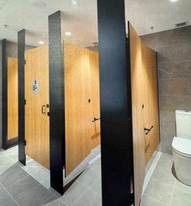 Modern public restroom with wooden partition doors and gray tile flooring. One stall door is slightly ajar, revealing an accessible stall equipped with a grab bar and toilet.