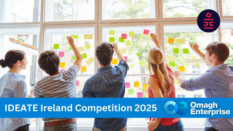 Five people facing a large window are sticking colorful sticky notes on the glass. A banner at the bottom reads, IDEATE Ireland Competition 2025 with the Omagh Enterprise logo on the right.