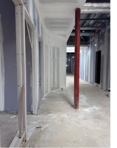 Construction site hallway with unfinished walls and exposed ceiling. A red support beam stands in the middle, surrounded by drywall panels. The floor is covered in dust, and wires hang from the ceiling, indicating ongoing construction work.