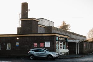 The old Health Centre building with a car parked in front. The building has a chimney and a sign that reads pharmacy on the facade. There are red and white signs near the entrance, and a canopy covers the doorway. Trees and a pale sky are in the background.