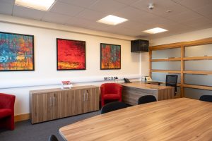 Office room with a large wooden table surrounded by black chairs. Colorful abstract art decorates the wall, and two red chairs are near a wooden cabinet. A phone and papers are on the desk. Nearby, there are shelves with glass panels.