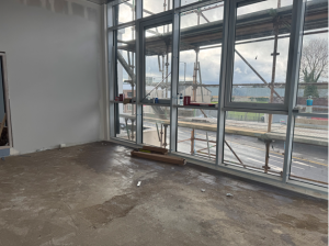 Unfinished room with large windows overlooking scaffolding and a cloudy sky. The floor appears bare and the walls are partially painted. Some tools and construction materials are near the window.