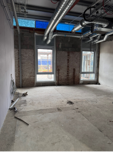 An unfinished room with exposed ducts and partially constructed walls. Two large windows reveal a glimpse of the exterior. The floor is bare concrete, and some construction materials and wires are scattered around.