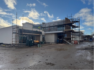 The old Health Centre transforming into a modern digi-hub building under construction with scaffolding on a muddy site. The structure features large glass windows and white walls. The sky is partly cloudy, and construction equipment is visible around the site.