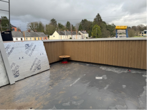 A rooftop, looking across Old Mountjoy Road. with a gray surface and a small round red table. Insulation panels lean against a wooden railing. In the background, there are houses, trees, and a partial view of construction equipment under a cloudy sky.