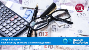 A calculator, glasses, and pens rest on British banknotes, with a banner inviting Omagh businesses to comment on future minimum wage rates.