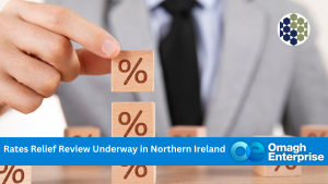 A person in a suit stacks wooden blocks with percent signs, illustrating a rates relief review in Northern Ireland. Omagh Enterprise logo is displayed on a blue banner.