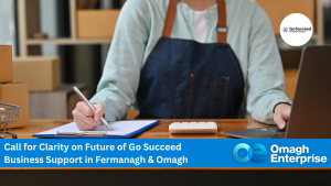 A person sits at a desk with a clipboard, pen, and calculator, surrounded by cardboard boxes. A banner reads: Call for Clarity on Future of Go Succeed Business Support in Fermanagh & Omagh, Omagh Enterprise.
