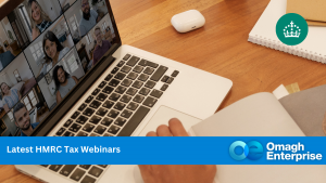 Person taking notes during a virtual meeting on a laptop, with coffee cups, a notebook, and Omagh Enterprise branding visible on the table.
