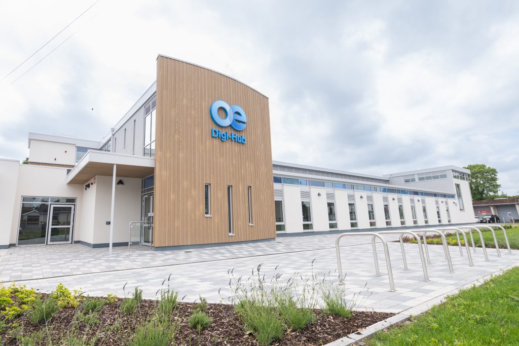 Modern Omagh Enterprise Digi-Hub building with a "Digi-Hub" sign on a wooden facade, surrounded by landscaping, bike racks, and a paved walkway under a cloudy sky.