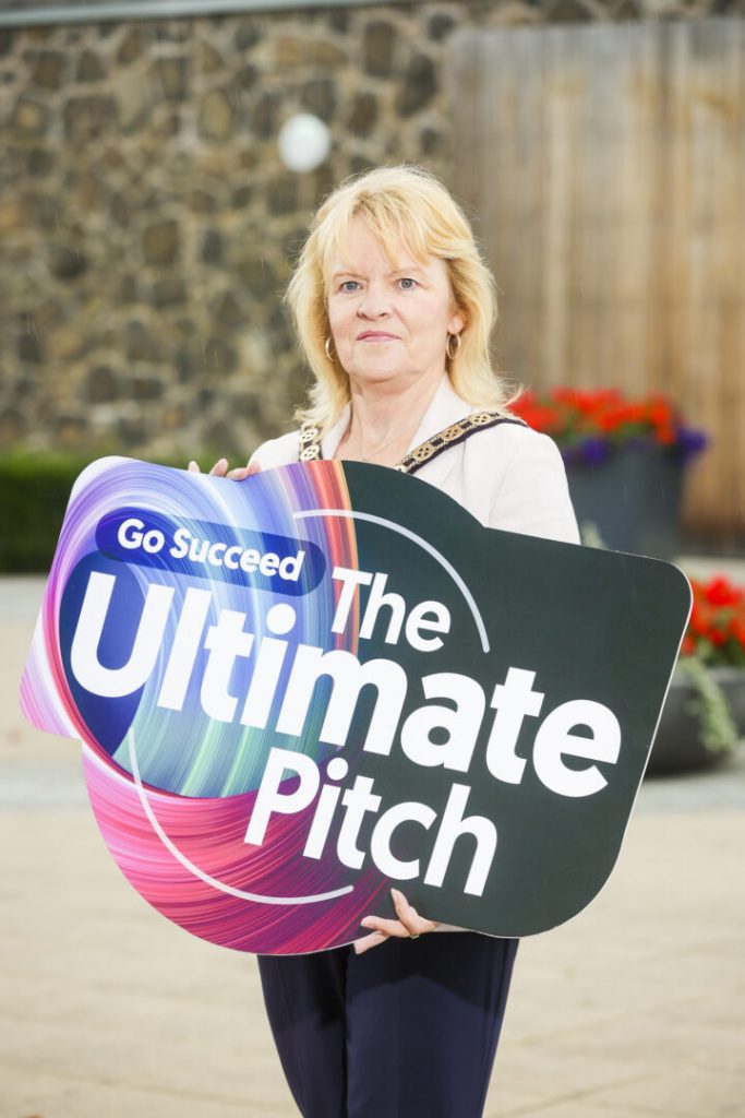 Councillor Shirley Hawkes, Vice Chair of Fermanagh and Omagh District Council stands outdoors, holding a large colorful sign that reads “Go Succeed The Ultimate Pitch” against a blurred background with stone walls and flowers.