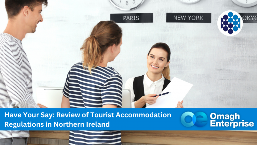 A hotel receptionist hands documents to two guests at a front desk, with city clocks on the wall and an Omagh Enterprise banner promoting accommodation regulation review.