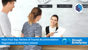 A hotel receptionist hands documents to two guests at a front desk, with city clocks on the wall and an Omagh Enterprise banner promoting accommodation regulation review.