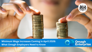 A person holds two uneven stacks of coins, with a banner about minimum wage increases for Omagh employers in April 2026 at the bottom.