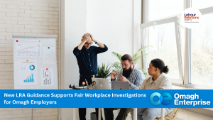 Three people sit around a desk with laptops and papers, discussing work in a bright office. One person stands, holding their head in frustration. Charts and graphs are displayed on the wall. A banner reads, Omagh Enterprise.