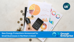 A calculator, coins, banknotes, and business charts on a desk, with a banner announcing new energy protections for small businesses in Northern Ireland from Omagh Enterprise.