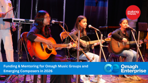 Three young women play acoustic guitars and sing into microphones on stage during a live music event, with banners promoting Omagh Enterprise and music funding programs.