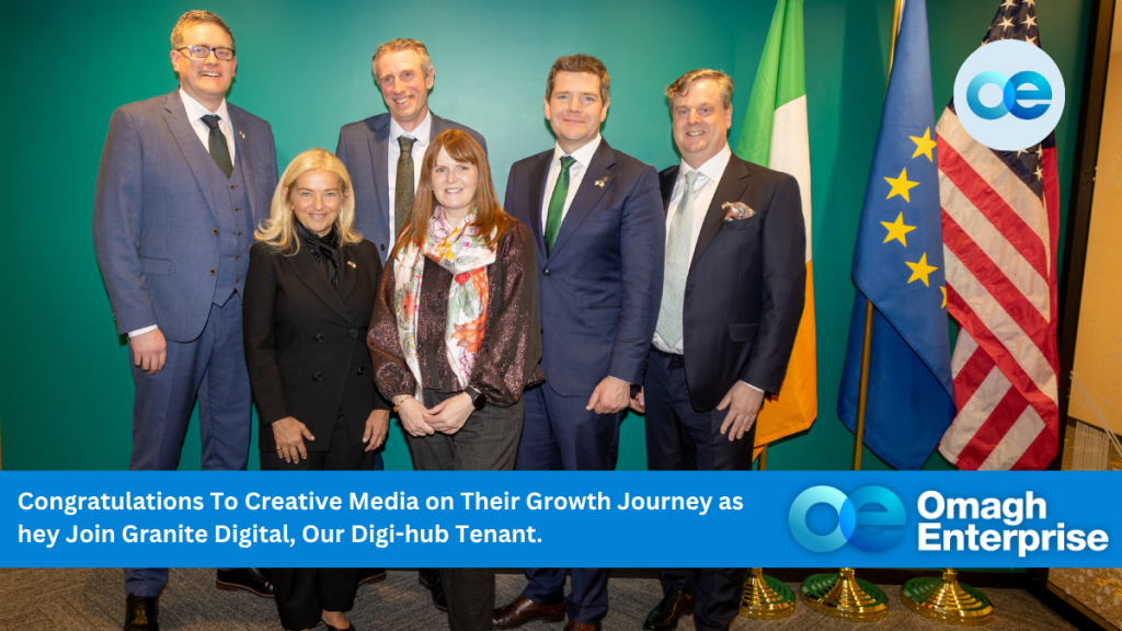 Six professionally dressed people pose for a group photo indoors beside the Irish and EU flags, with a congratulatory banner for Creative Media and Omagh Enterprise branding. In the photo are: Northern Ireland Economy Minister Caoimhe Archibald, Irish Enterprise Minister Peter Burke. Creative Media's Marcus Isherwood and Granite's Chief Executive Conor Buckley