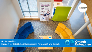 Overhead view of a modern office lounge with blue at Omagh Enterprise in Gortrush, yellow, and green seating, a coffee table, and a business support banner. “Omagh Enterprise” and “Go Succeed NI” logos are visible.