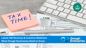 A desk with tax forms, a keyboard, a silver pen, and a pink sticky note reading "TAX TIME!" alongside a banner for an Omagh Enterprise tax webinar.