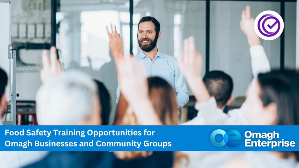 A man stands at the front of a classroom, speaking to attendees with raised hands. A banner promotes food safety training for Omagh businesses and community groups by Omagh Enterprise.