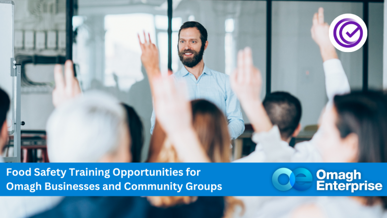 A man stands at the front of a classroom, speaking to attendees with raised hands. A banner promotes food safety training for Omagh businesses and community groups by Omagh Enterprise.
