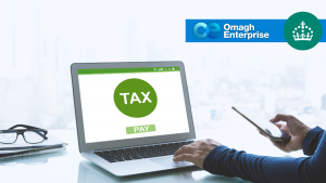A person uses a laptop displaying a green TAX button and PAY option, while holding a smartphone. The Omagh Enterprise logo and a crown symbol appear in the top right corner.