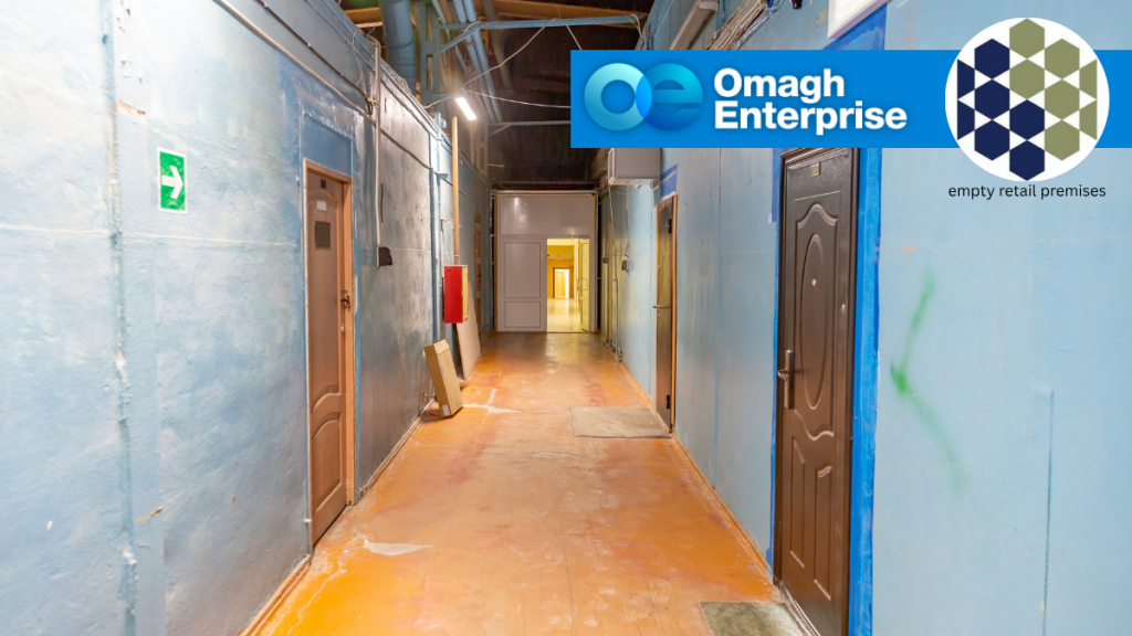 A dimly lit hallway with blue walls, several closed doors, and exposed pipes. The Omagh Enterprise and Empty Retail Premises logos appear in the upper right corner.
