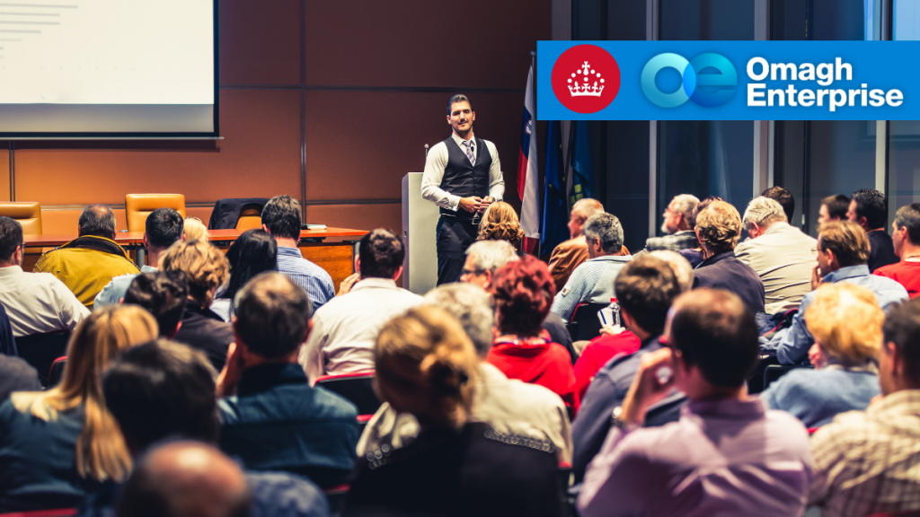 A man stands and speaks to an audience at a conference or seminar. The Omagh Enterprise logo is displayed in the upper right corner.