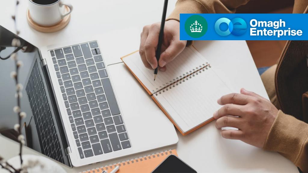 A person writes in a notebook beside a laptop and coffee cup; Omagh Enterprise and UK government logos appear in the top right corner.