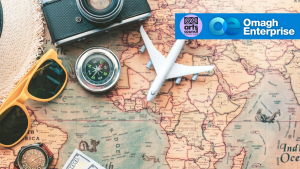 A vintage camera, compass, toy airplane, sunglasses, and cash are arranged on a world map. The logos of the Arts Council and Omagh Enterprise are displayed in the top right corner.