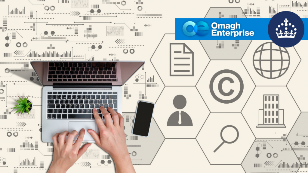 Hands typing on a laptop next to a smartphone and plant, with Omagh Enterprise and crown logos, and icons representing business concepts in the background.