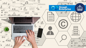 Hands typing on a laptop next to a smartphone and plant, with Omagh Enterprise and crown logos, and icons representing business concepts in the background.