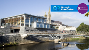 The Strule Arts and bridge over the river in front, set against older stone buildings and a church spire. Logos for Omagh Enterprise and Fermanagh & Omagh District Council are visible in the sky area.