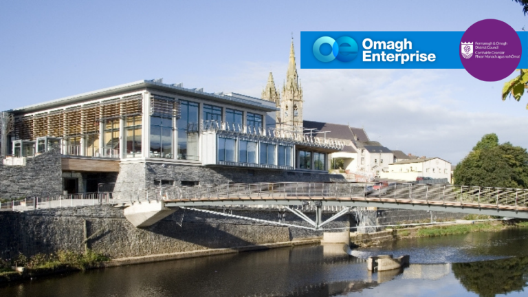 The Strule Arts and bridge over the river in front, set against older stone buildings and a church spire. Logos for Omagh Enterprise and Fermanagh & Omagh District Council are visible in the sky area.